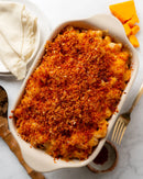 A baked macaroni casserole topped with a golden-brown breadcrumb crust, served in a white rectangular baking dish on a light marble surface, with folded napkins, a fork, and cheese cubes nearby.