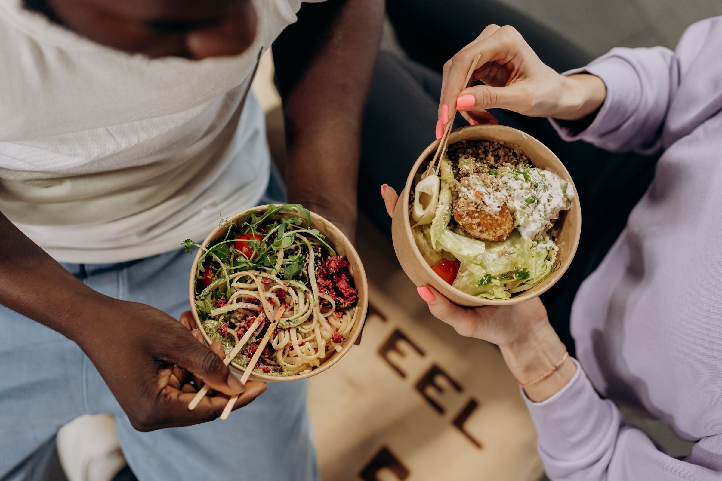 Two people holding paper bowls of food in a cafe setting. The person on the left holds a bowl with pasta, greens, and red toppings, while the person on the right holds a bowl with a fried item, greens, and white sauce.